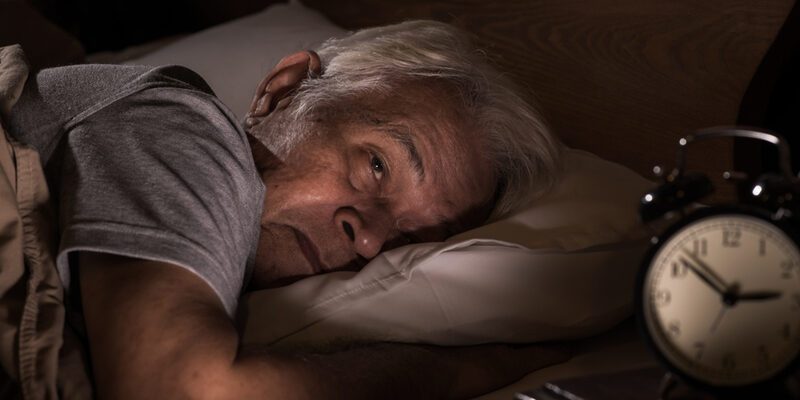 An image illustrating insomnia: a senior man resting in bed while an alarm clock reminds of the difficulties in achieving a restful sleep