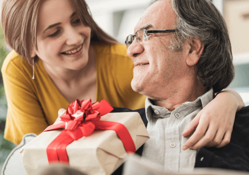 A woman holds a gift box while seated next to a man. The image depicts a scene related to home care